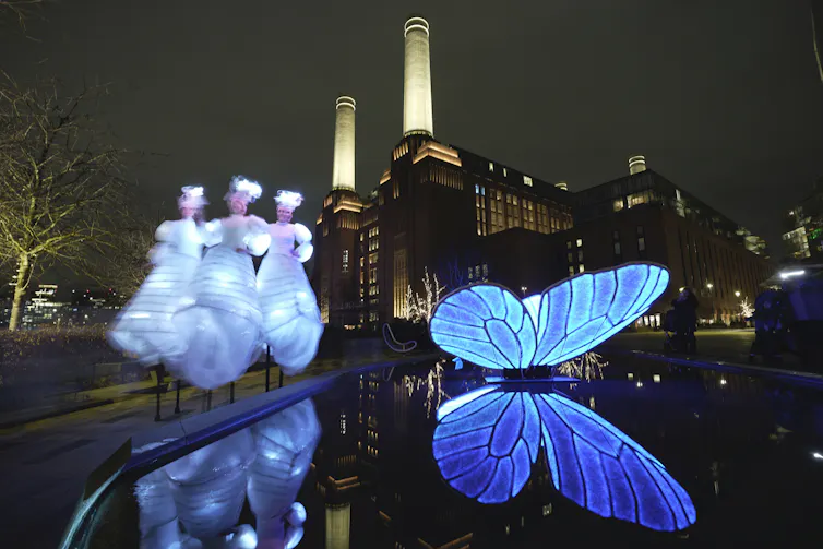 A blue illuminated butterfly art installation in front of an art deco decommissioned power plant at night time.