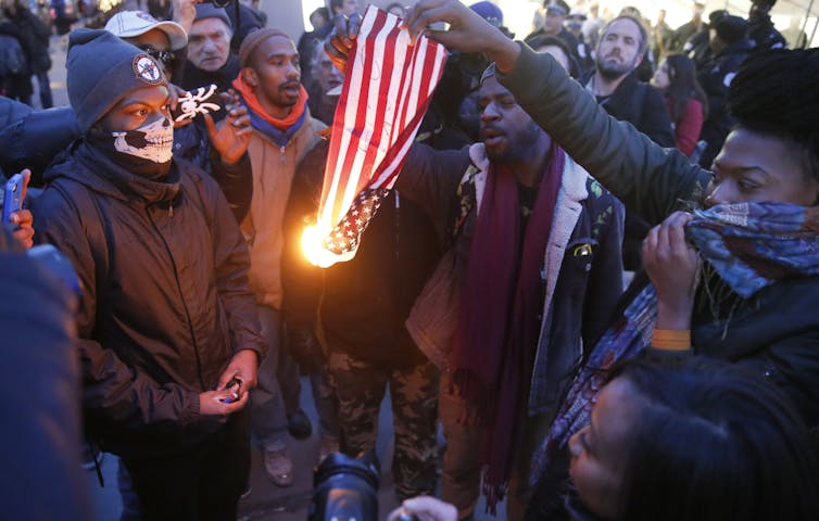 A group of people surround a U.S. flag that has been lit on fire.