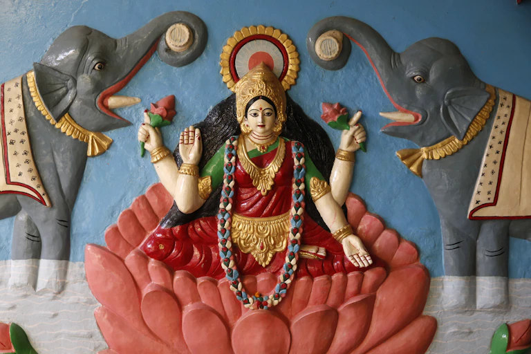 A sculpture showing a goddess in a red saree, seated on a lotus, which is flanked by two elephants.