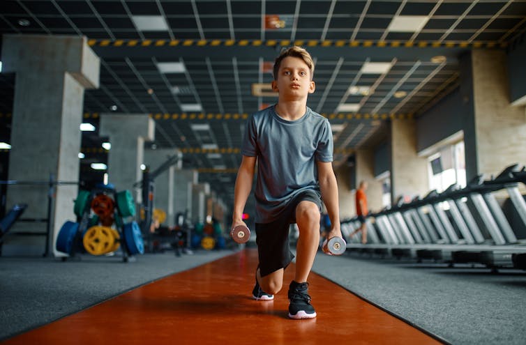 A boy does weighted lunges in a gym.