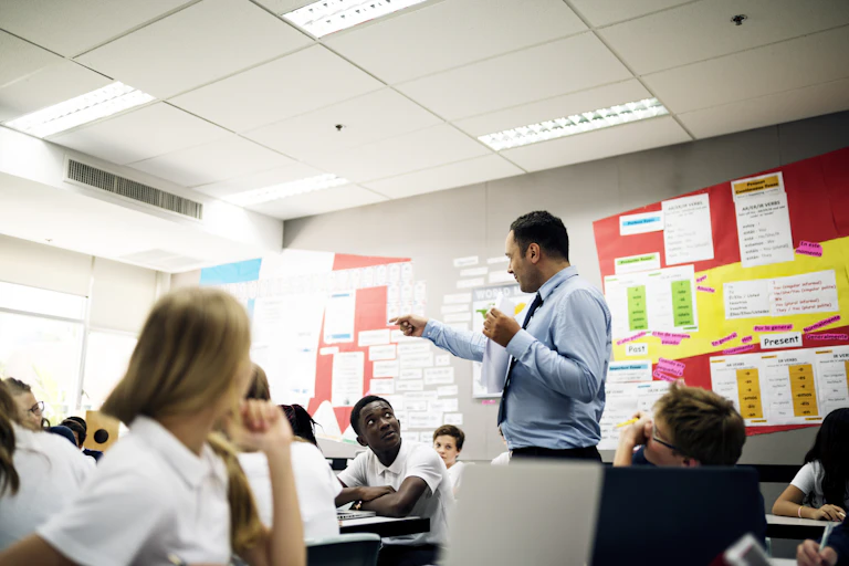 A teacher in shirt and tie stands in the middle of students, seated at desks. The teacher points to the left.
