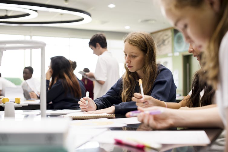 Students sit together at a desk in a classroom with pens and notebooks