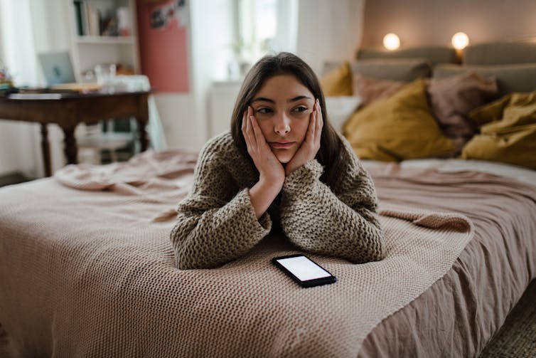 A teenage girl lies on a bed with her bands in her face. A phone is next to her on the bed.
