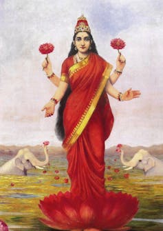 Panting showing a goddess in a red saree. She is standing on a blooming lotus, with two of her four hands holding the flower. She is flanked by two white elephants on either side.