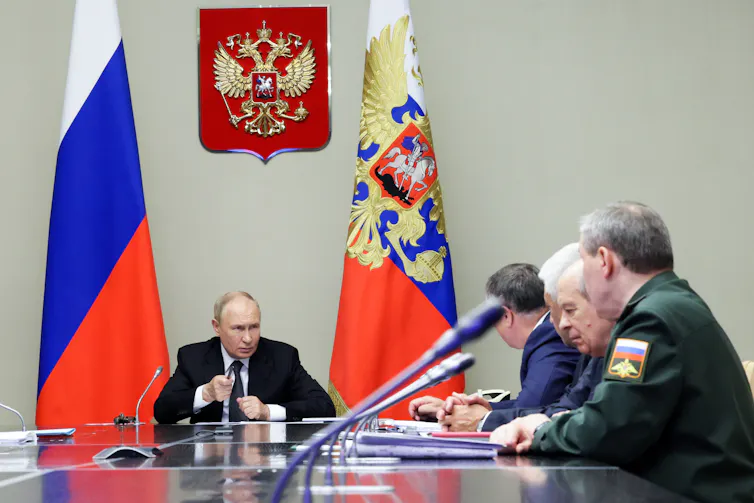 Vladimir Putin sits at one end of a long table, flanked by national flags, with members of the national security council facing him.