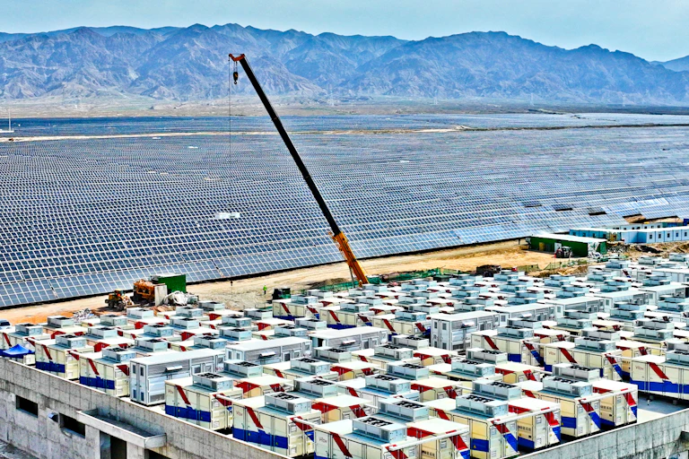 Hundreds of rows of solar panels on a sandy area next to dozens of giant batteries the size of shipping containers.
