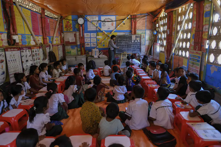 School children sit in a classroom as a teacher stands in front next to a blackboard.