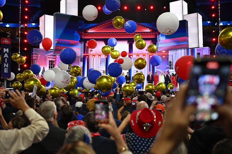 Red, white, blue and gold balloons cascading through the air in a large, crowded convention center.