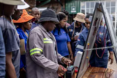 A group of people stand around a board with solar parts mounted on it, testing the different components as part of their training to become solar energy technicians
