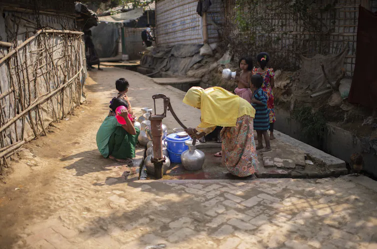 A woman in a yellow headscarf pumps water from a well into a metal container as children surround her.