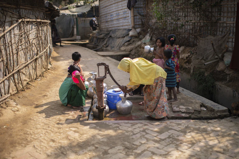 A woman in a yellow headscarf pumps water from a well into a metal container as children surround her.