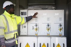 A man points to a big white box full of electrical components that are part of a solar energy system, telling people how it works