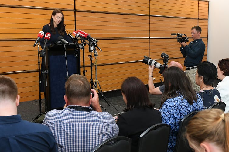 A woman with dark hair stands at a podium, surrounded by microphones and people with cameras crouching down in front of her.