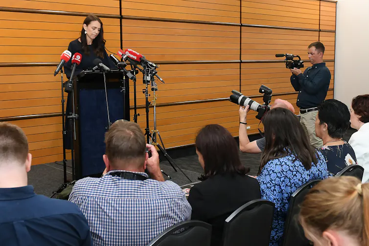 A woman with dark hair stands at a podium, surrounded by microphones and people with cameras crouching down in front of her.