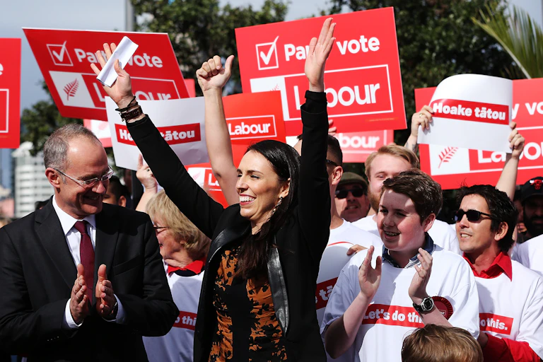 A white woman with dark hair raises her hands in the air, while people stand around her and hold red and white posters that say 'Labour' and 'Party vote' and have white tee-shirts that say 'let's do this.'