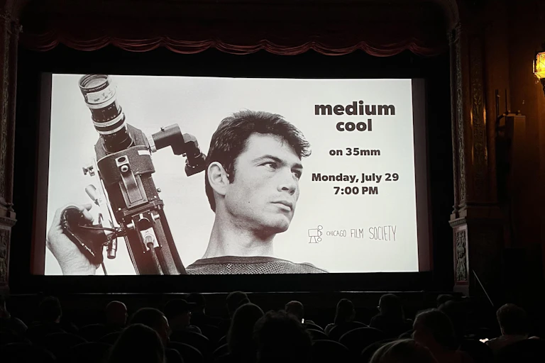 A dark haired man with a film camera in his right hand, on a promotion for a moved called 'Medium Cool.'