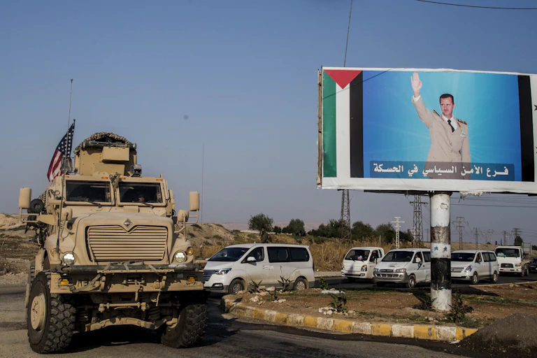 An armored vehicle flies tha American flag as it passes a billboard with a man saluting.