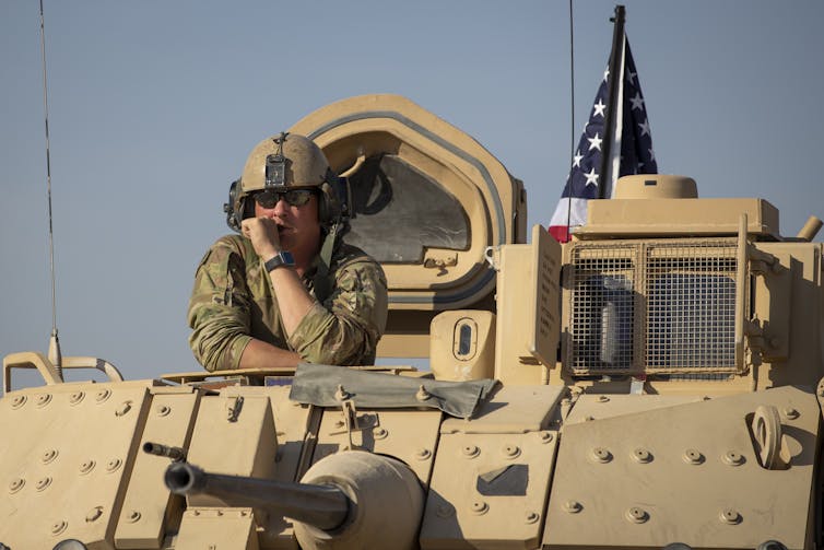 A man in army fatigues looks out from an armored vehicle.
