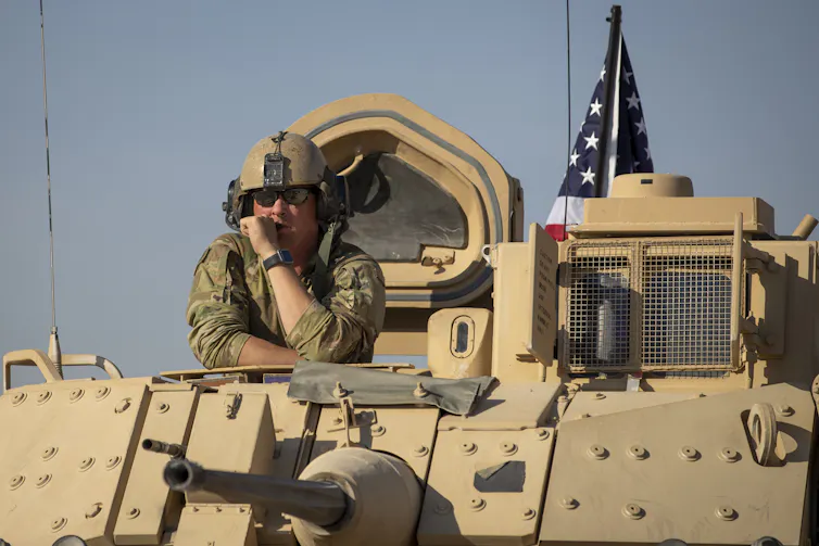 A man in army fatigues looks out from an armored vehicle.