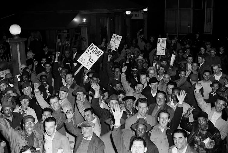 A large group of smiling men from a mix of races and ethnic backgrounds cheer and holds signs.