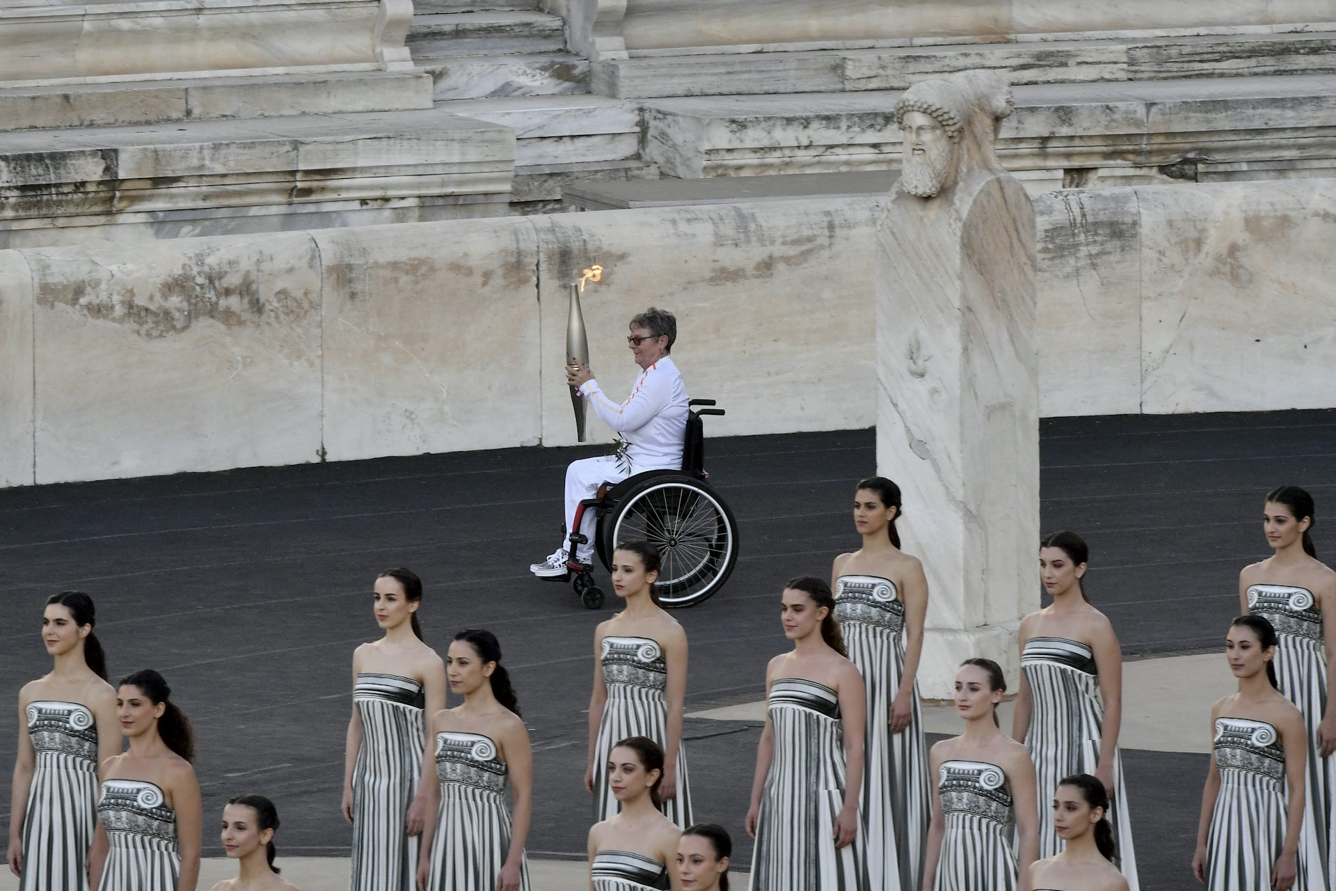 A white, older woman in a wheelchair carries a lit torch across the floor of a stadium