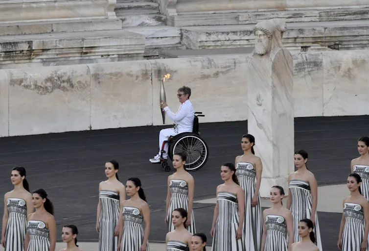 A white, older woman in a wheelchair carries a lit torch across the floor of a stadium