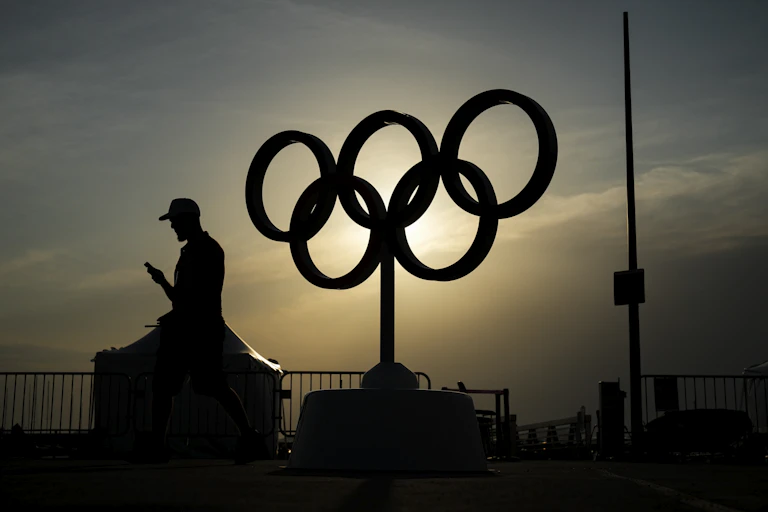 The sun sets behind a statue of the Olympic rings, silhouetting them and a man walking past