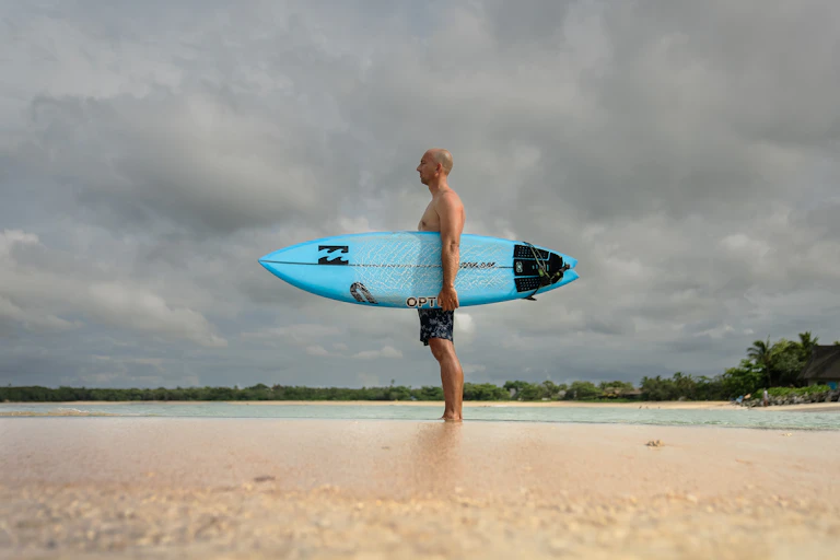 A man stands on the sand holding a surf board.