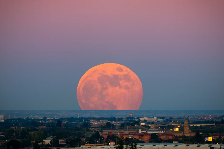A seemingly enormous orange Moon rising over a city at dusk.