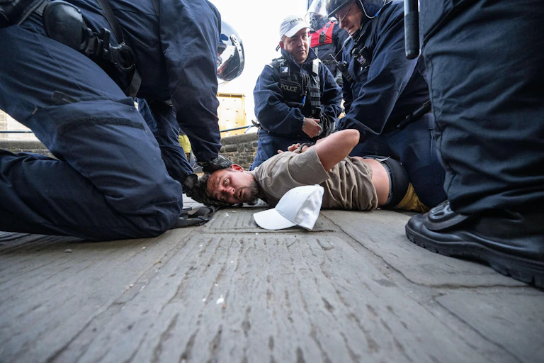 A man lying face down on the pavement being pinned down and handcuffed by riot police.