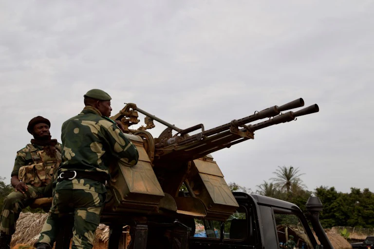 Two men wearing military fatigues in the back of a military vehicle kitted with canons