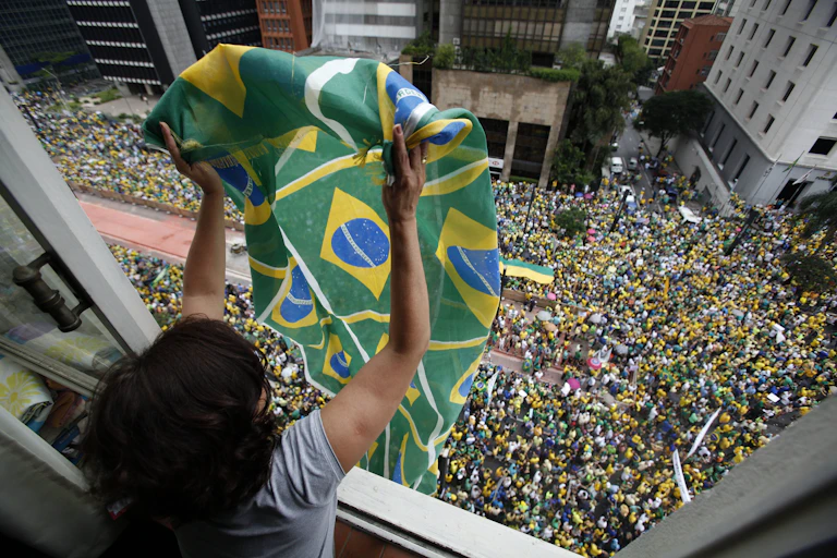 A woman waving a Brazilian flag out of her window as thousands of protesters gather on the street below.