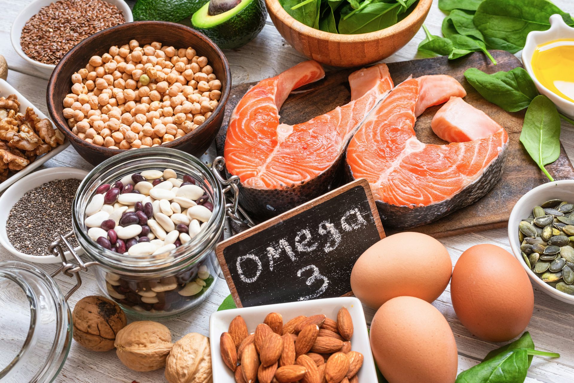 Une tranche de saumon, des œufs, des légumineuses, des amandes, des épinards, etc. sont posés sur une table. Au milieu, est posée une ardoise sur laquelle est écrit Oméga 3