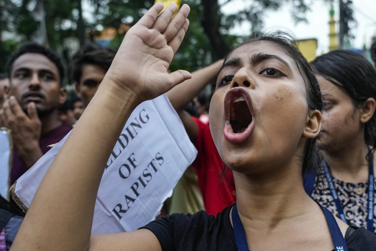 A woman in the foreground of a protest, holds her hand in the air and opens her mouth wide to yell.