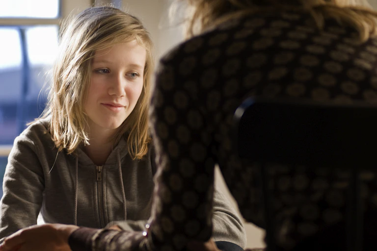 A teen girl looks sad as she speaks with a counselor in an office.
