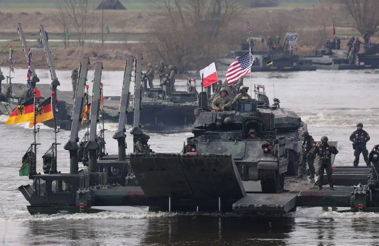 Military vehicles flying U.S., Polish, German and U.K. flags drive across a river.