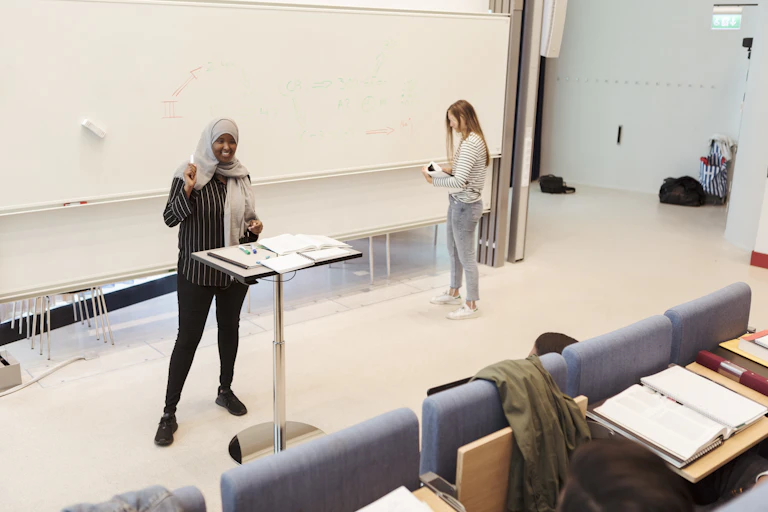 A female student stands at a lectern while another stands at a whiteboard.