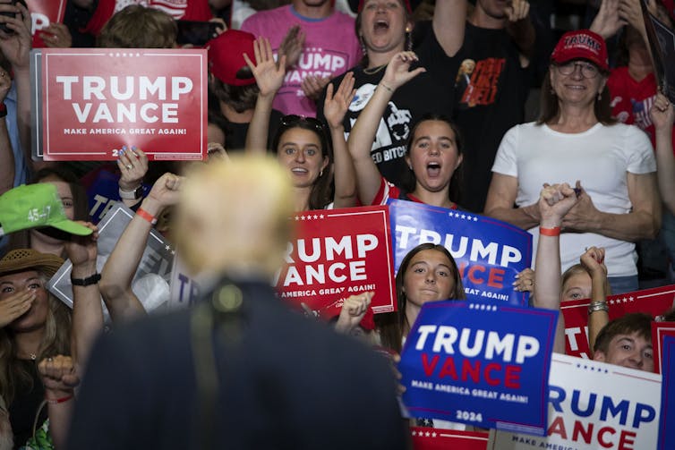 People hold posters that say 'Trump Vance' and face toward a person wearing a dark suit who is blurry