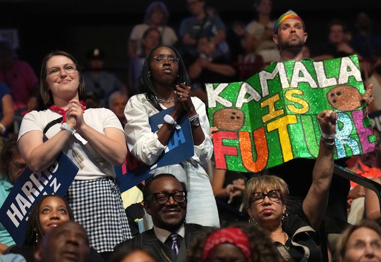 Three young people stand and two of them, women, hold their hands against their chests. One person holds a poster that says 'Kamala is future.'