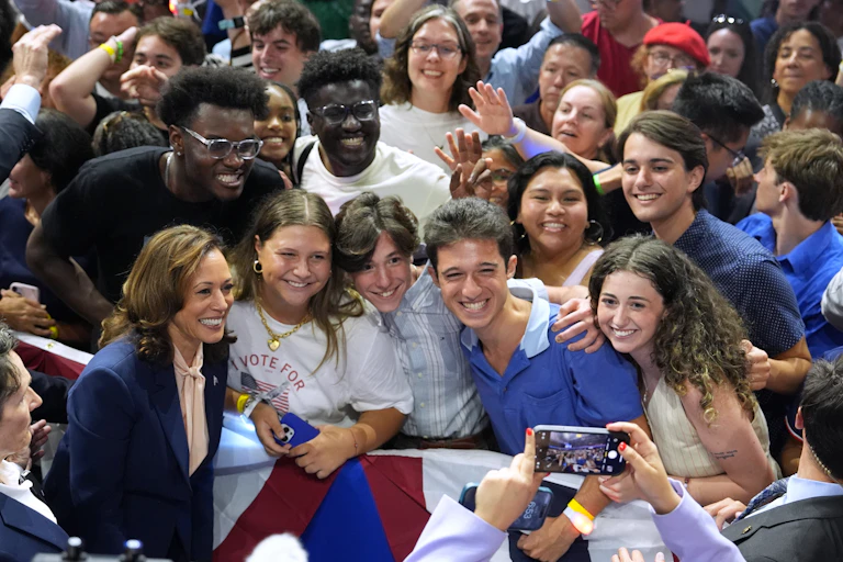 A woman wearing a pantsuit smiles next to a group of four young people, who are surrounded by other young people, all smiling.