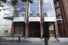 People walk by a glass and brick-fronted building with the name 'Pierre de Coubertin stadium' on it