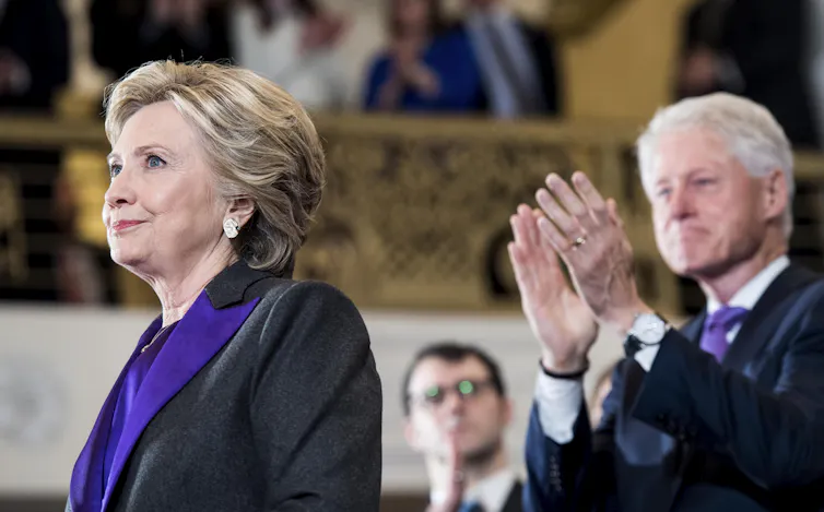 A resigned looking woman in a dark jacket and purple shirt is applauded by a white haired man behind her.