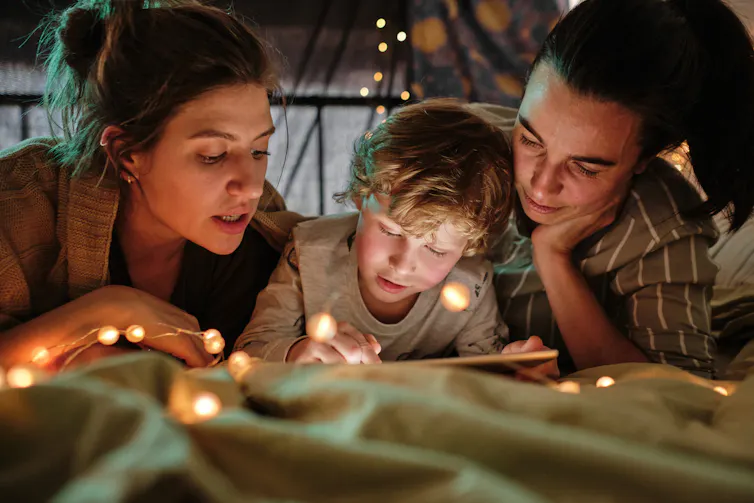 Two mums and a young child read together. They are surrounded by fairy lights.
