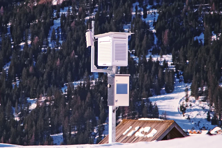 Weather equipment housed within a white box atop a pole in snowy mountains.
