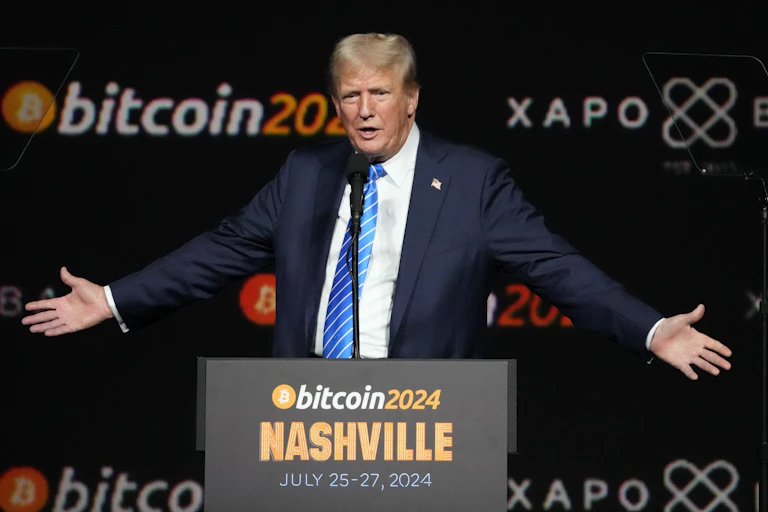 A man with blond-grey hair stands behind a lectern with his arms spread. a bitcoin2024 banner is behind him.