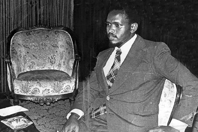 A man wearing a suit sits in a chair holding a cigarette in his right hand.