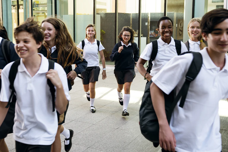Teenage school children in white polo shirts and dark jumpers and pants walk and run outside a building.