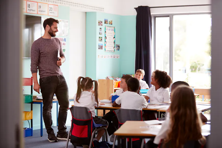A teacher stands and holds a tablet. Primary-aged children sit at desks, looking at the teacher.