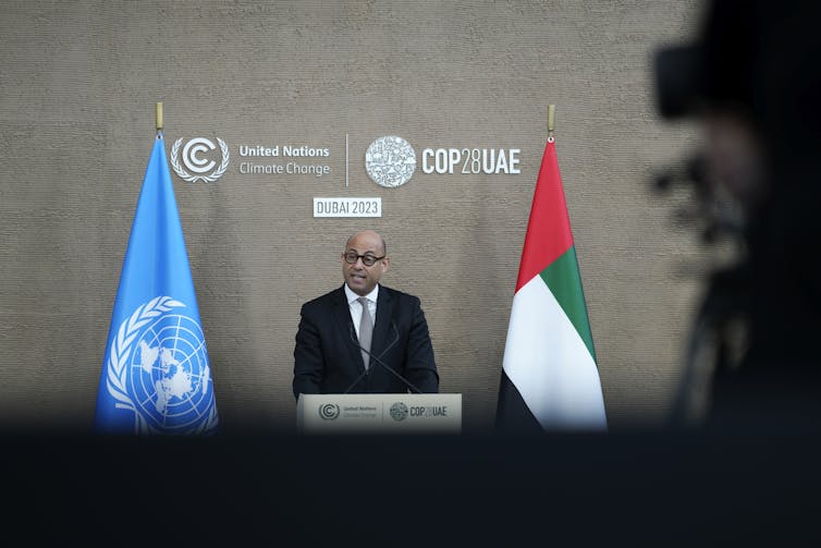 A man in a suit stands between two flags at a podium