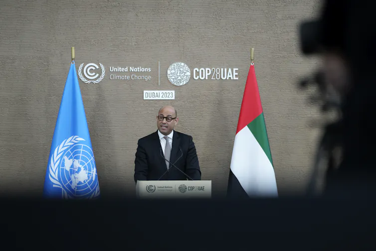 A man in a suit stands between two flags at a podium